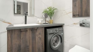 A modern bathroom with a marble wall, a wooden cabinet, a washing machine, a sink, a mirror, and a potted plant.