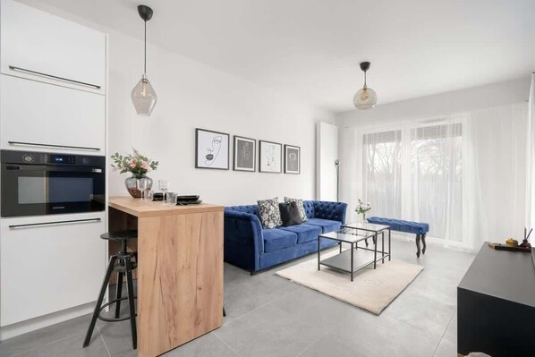 A modern kitchen with white cabinets and a built-in oven, a blue sofa with a coffee table, and a dining area with a black table and chairs.