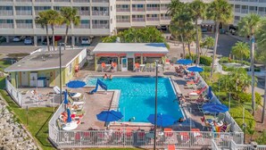 Et hotel med en swimmingpool omgivet af liggestole og parasoller, et overdækket område og en parkeringsplads.