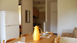 Un comedor con una mesa de madera dispuesta para dos personas, una cocina al fondo y una sala de estar con sofá.
