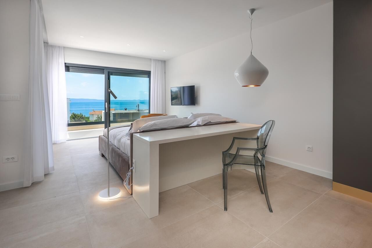 Un dormitorio moderno con una cama grande, un escritorio y una silla. Hay un televisor en la pared y vista al mar a través de la ventana.