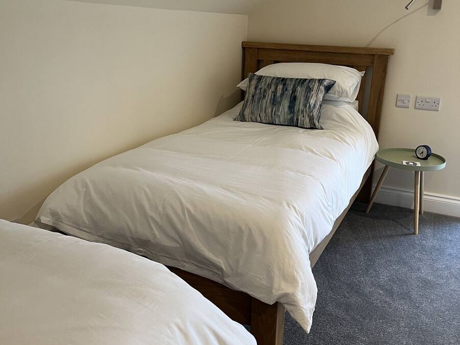 A single bed with white bedding in a room with a sloped ceiling and a small round table with a clock.