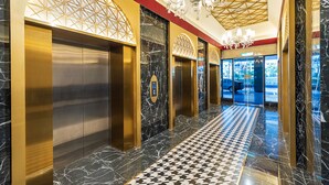 Un área de ascensor moderna con pisos de mármol y detalles dorados, que presenta un diseño geométrico en el techo y un candelabro.