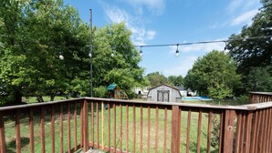 Una terraza de madera con vista a un patio trasero, que incluye una casa pequeña y una piscina.