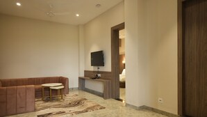 A modern living room with a brown sofa, a small round table, a flat-screen TV mounted on the wall, and a wooden bench. - Mansa Club And Resort (Ujjain)