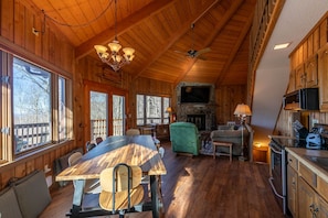 Interior de una cabaña de madera con una mesa de comedor, sillas, chimenea, televisor y zona de cocina.