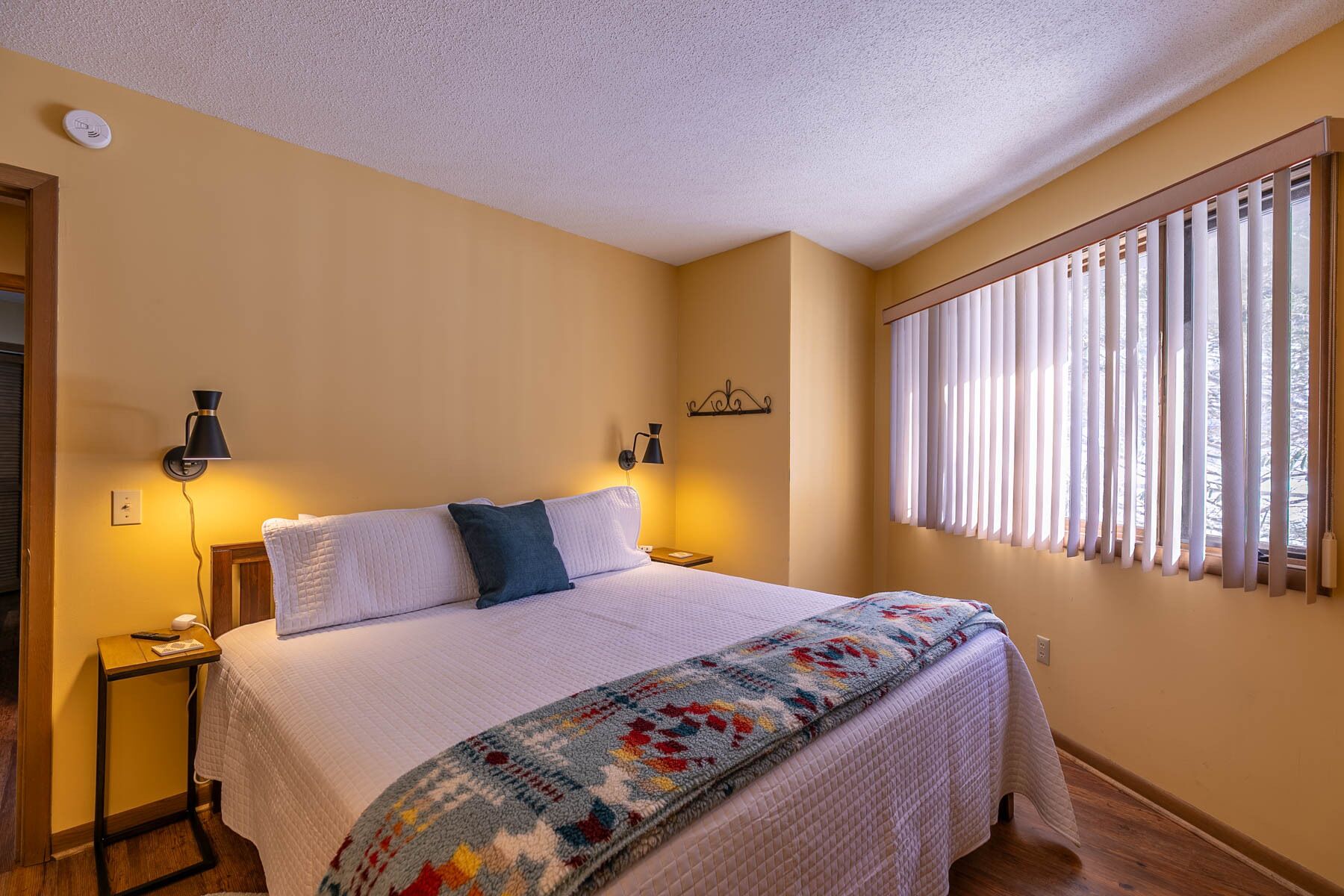 A bedroom with a bed, bedside lamps, a window with blinds, and a wall-mounted light fixture.