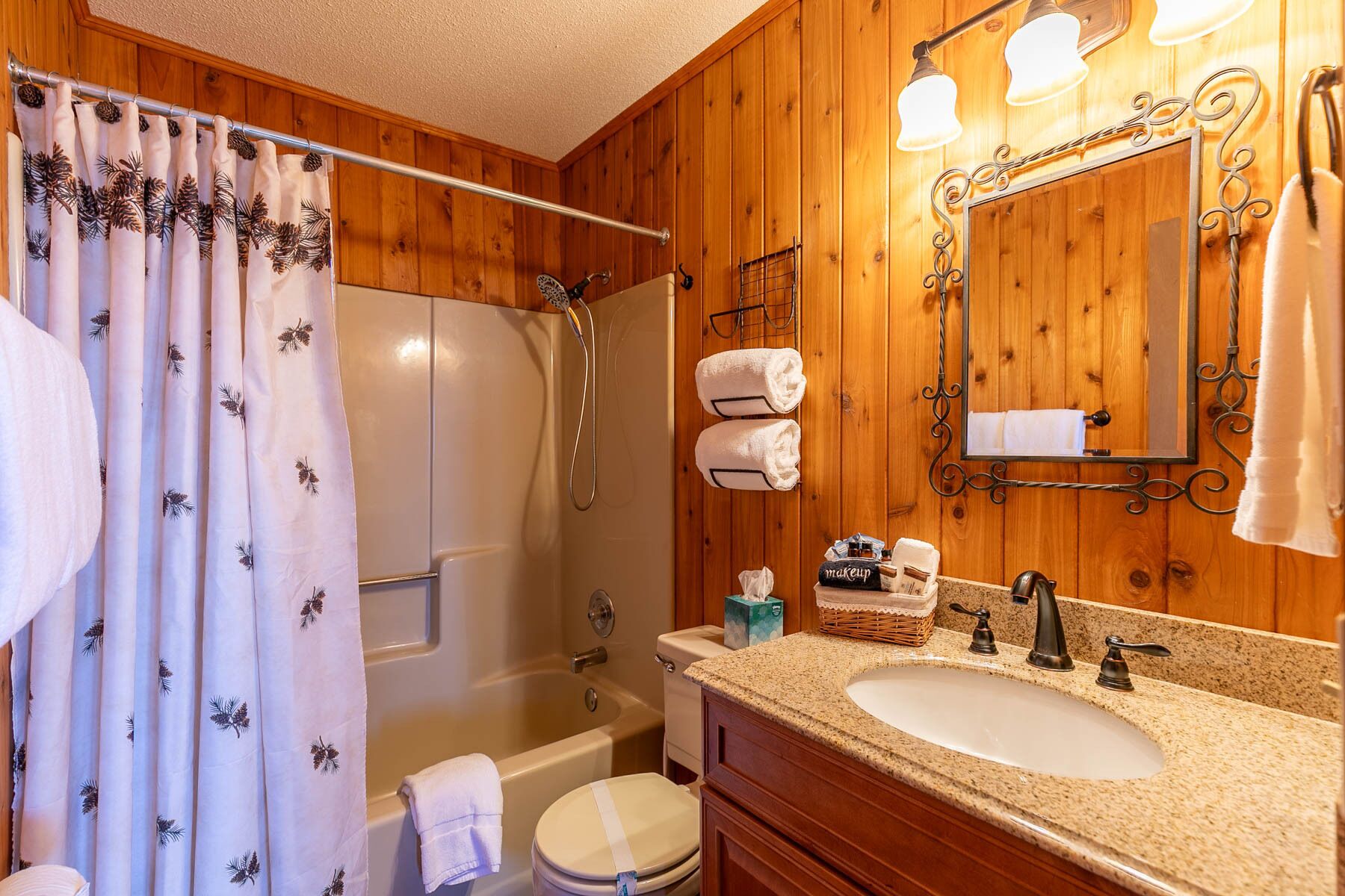 A bathroom with a wooden interior, a bathtub, a toilet, a sink with a mirror, and a shower curtain with a pine leaf pattern.