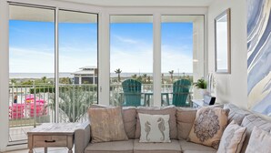 Un balcón con vista a la playa y palmeras, un sofá color beige con cojines decorativos, y una mesa con sillas.