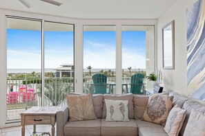 A balcony with a view of the beach and palm trees, a beige sofa with decorative pillows, and a table with chairs.