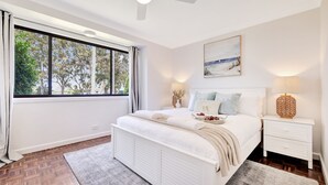 A bedroom with a large window, a bed with white bedding, a bedside table with a lamp, and a painting on the wall. - Plumeria – 350m to beach track, pet friendly (Callala Beach)