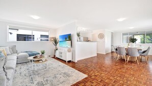 A modern living room with a sofa, coffee table, and TV. There's a kitchen area with a dining table and chairs. - Plumeria – 350m to beach track, pet friendly (Callala Beach)