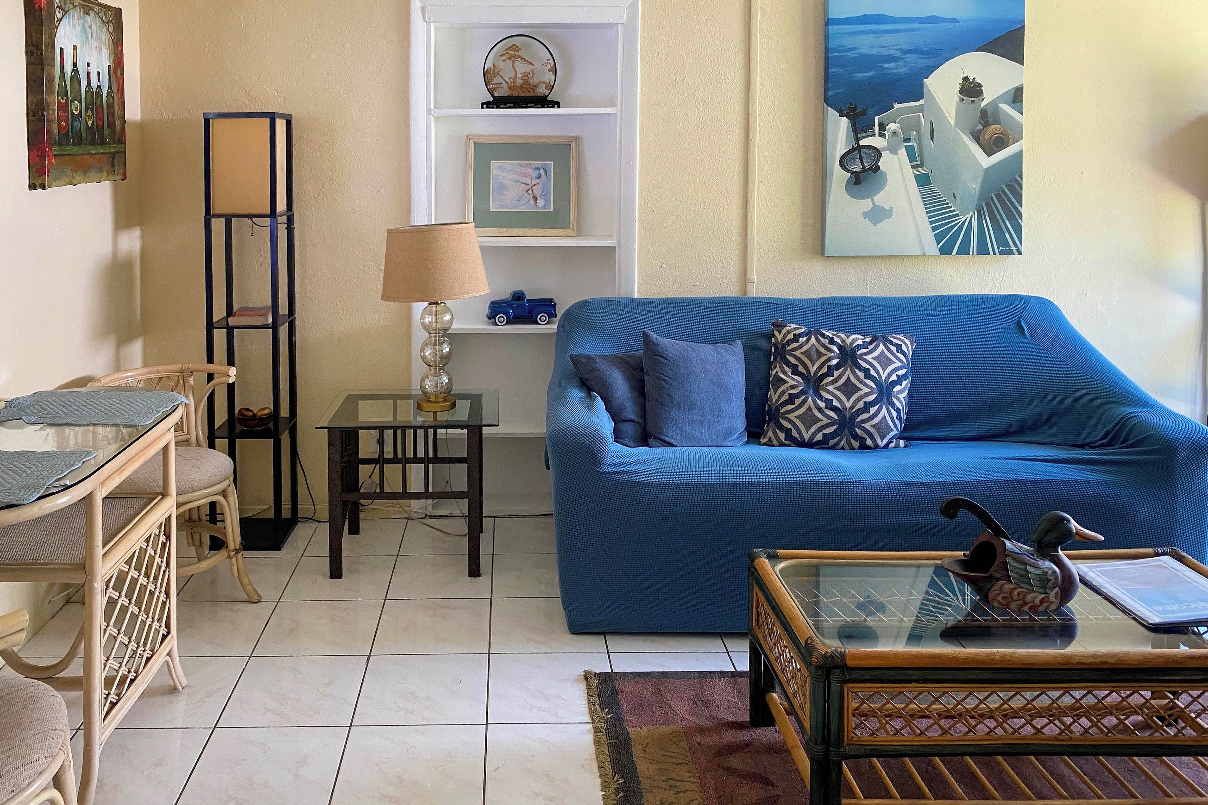 A living room with a blue sofa, a wooden coffee table, a wicker chair, a small table with a lamp, and a wall with framed artwork.