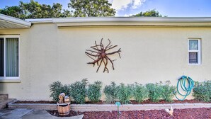 Una casa con un original mural de un árbol, un ventanal pequeño y un jardín con plantas y una manguera.