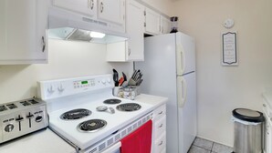 Una cucina con mobili bianchi, un fornello, un frigorifero e un cestino.