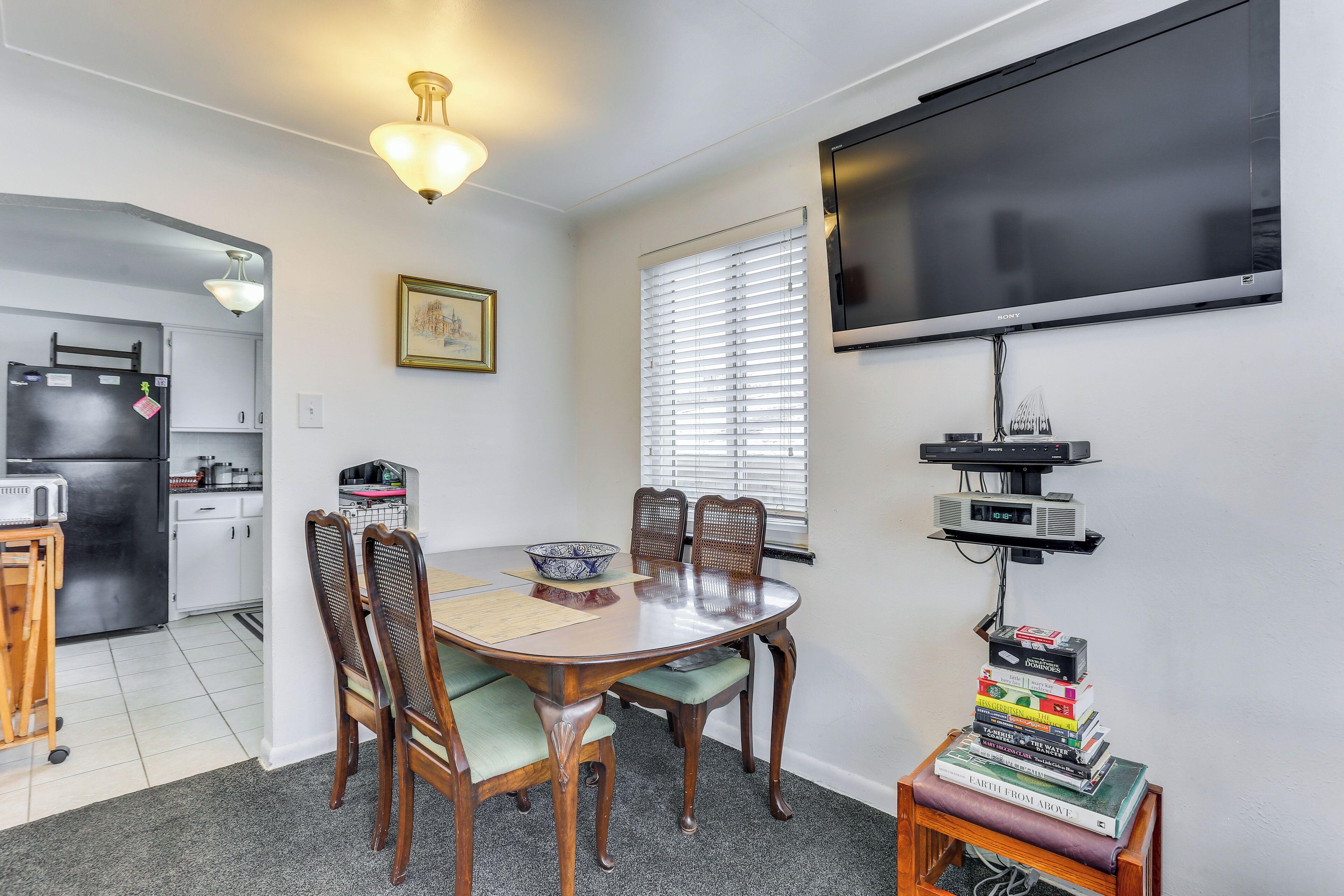 A dining area with a round table, chairs, a television mounted on the wall, and a kitchen in the background.