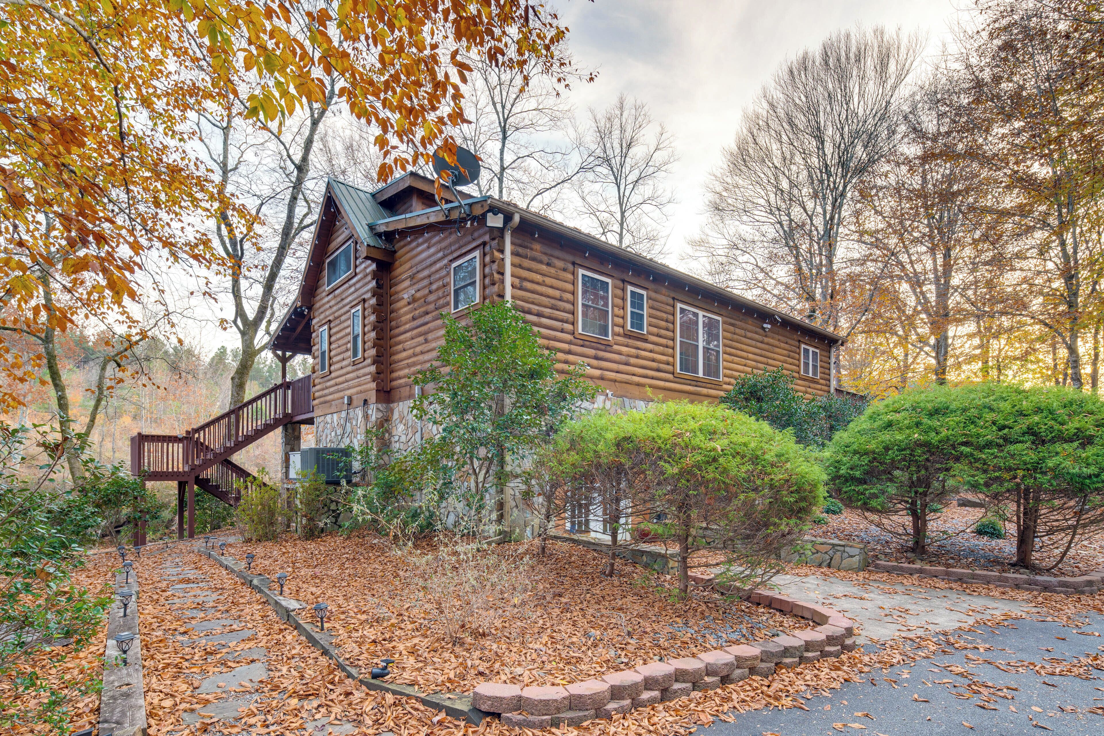 A log cabin with a wooden staircase leading to the entrance, surrounded by trees and a leaf-covered ground.