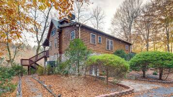 A log cabin with a wooden staircase leading to the entrance, surrounded by trees and a leaf-covered ground.