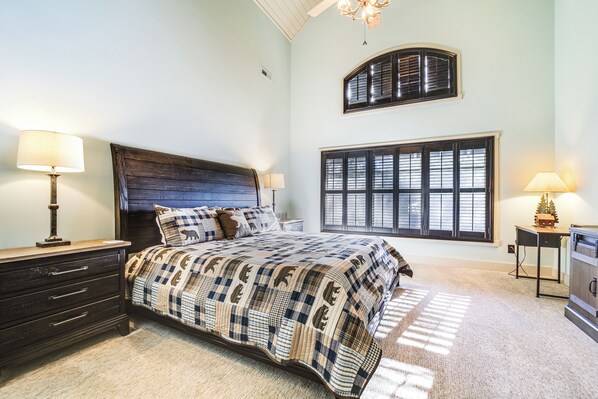 Un dormitorio con una cama grande, dos mesitas de noche con lámparas, un aparador, una ventana con contraventanas y un ventilador de techo.