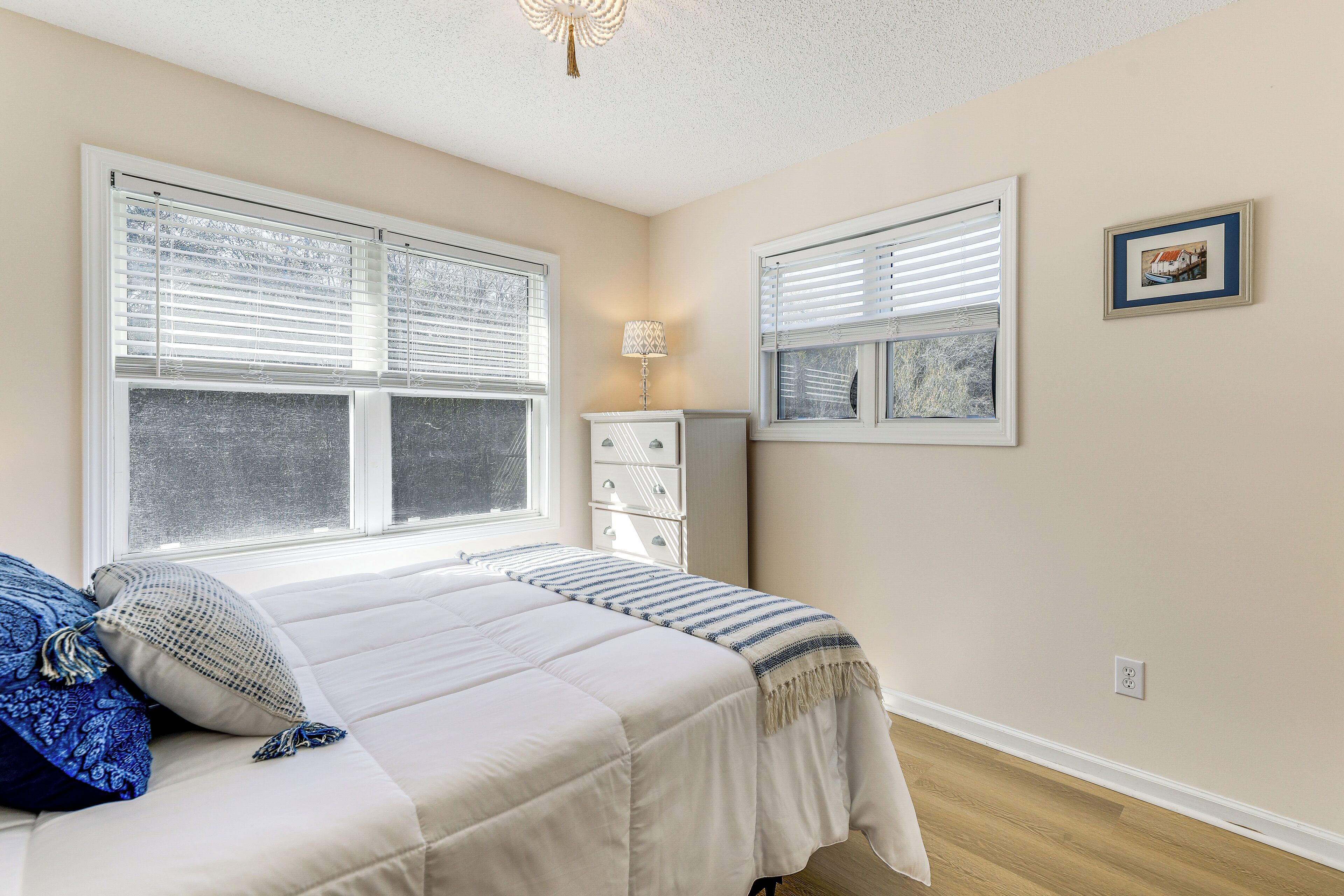 Une chambre à coucher avec un lit, deux fenêtres équipées de stores, une commode, une lampe et un tableau encadré accroché au mur.