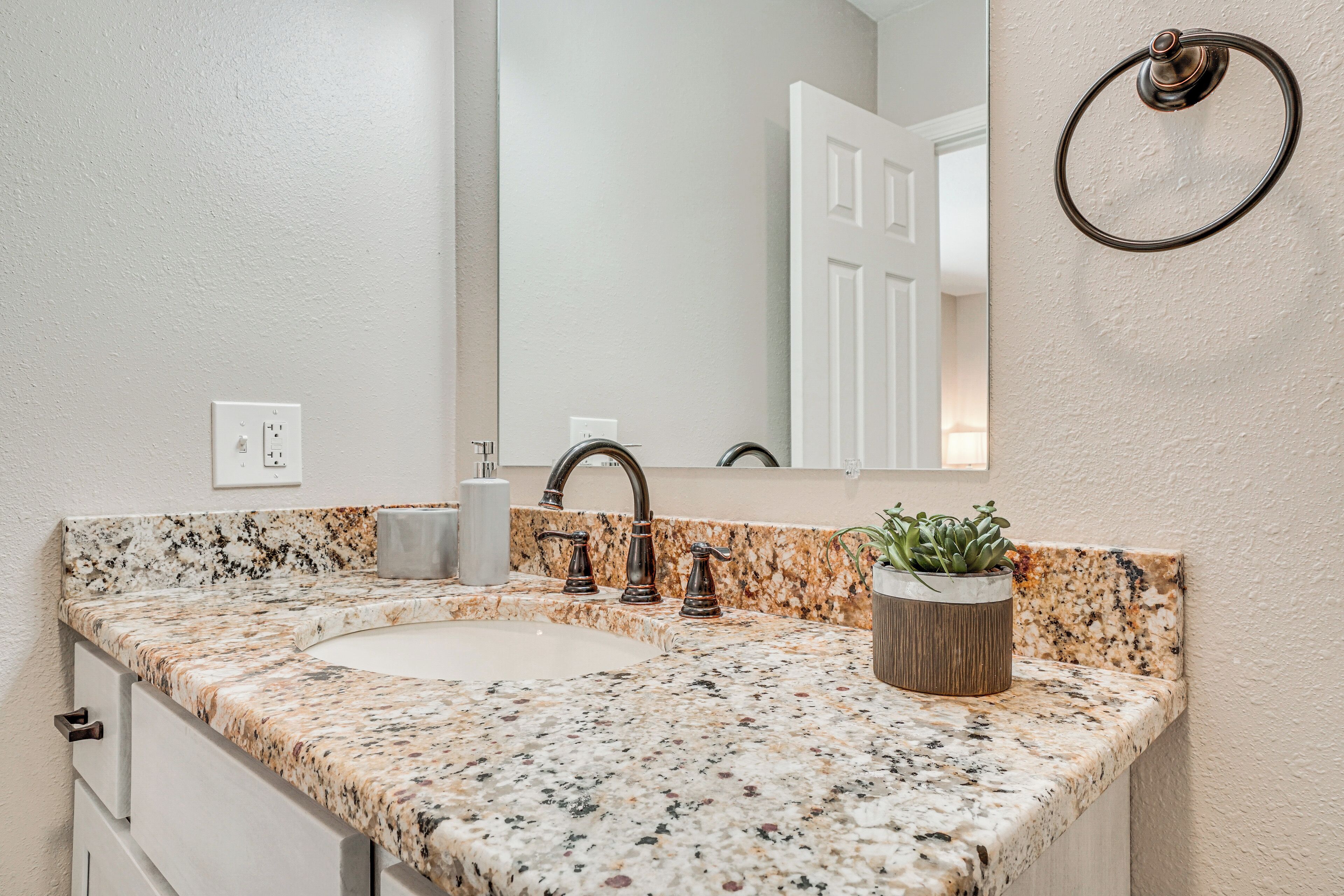 A bathroom with a granite countertop, a sink, a mirror, a towel ring, and a potted plant.