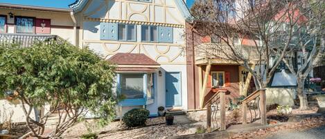 Una casa tradicional de dos pisos con un techo pronunciado, un porche delantero y un pequeño jardín con un árbol y plantas en macetas.