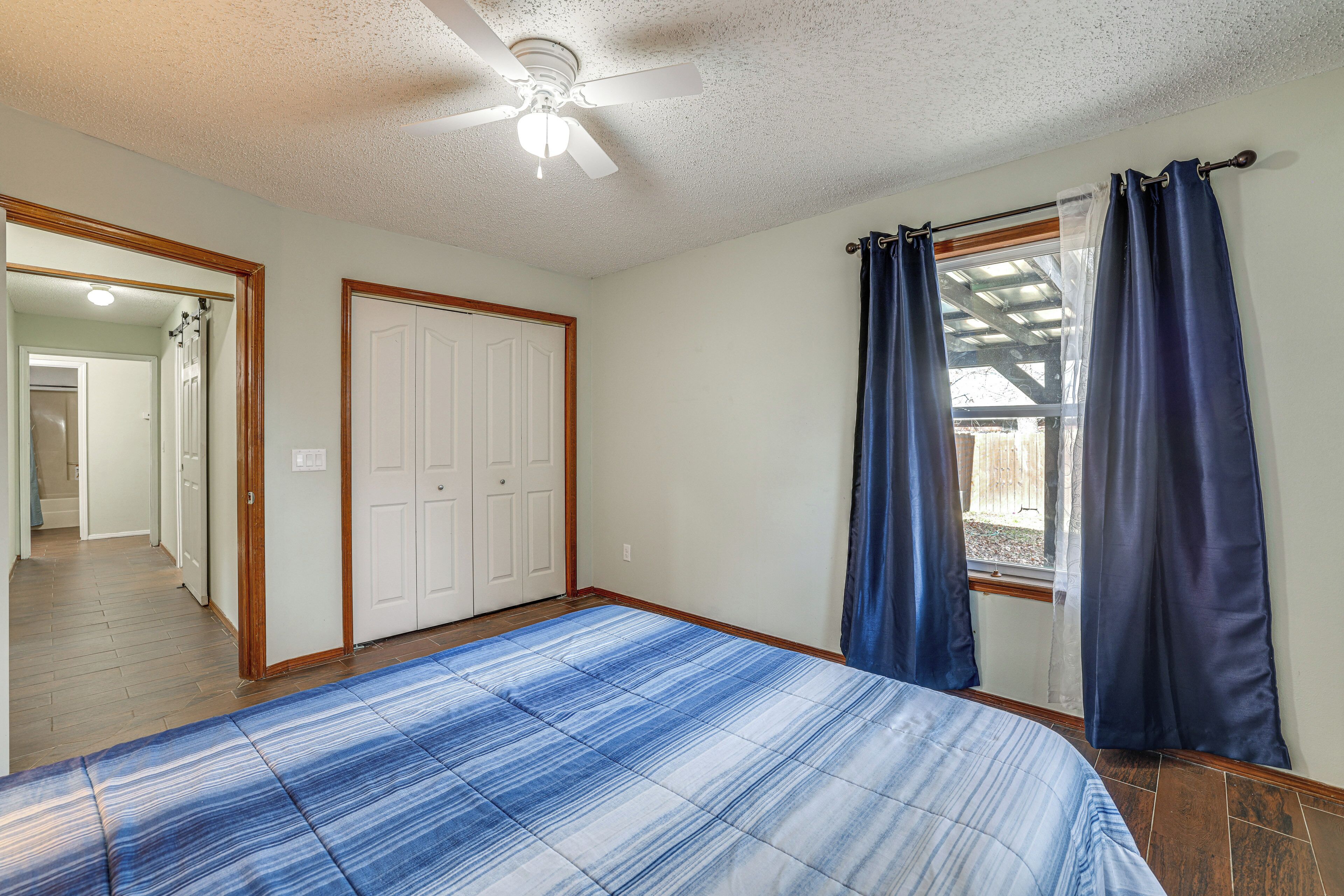 A bedroom with a bed, a ceiling fan, a window with curtains, and a door leading to a hallway.