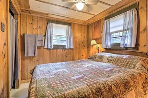 Une chambre à coucher aux murs lambrissés, avec un grand lit, deux fenêtres avec des rideaux, un ventilateur de plafond et une table de chevet avec une lampe.