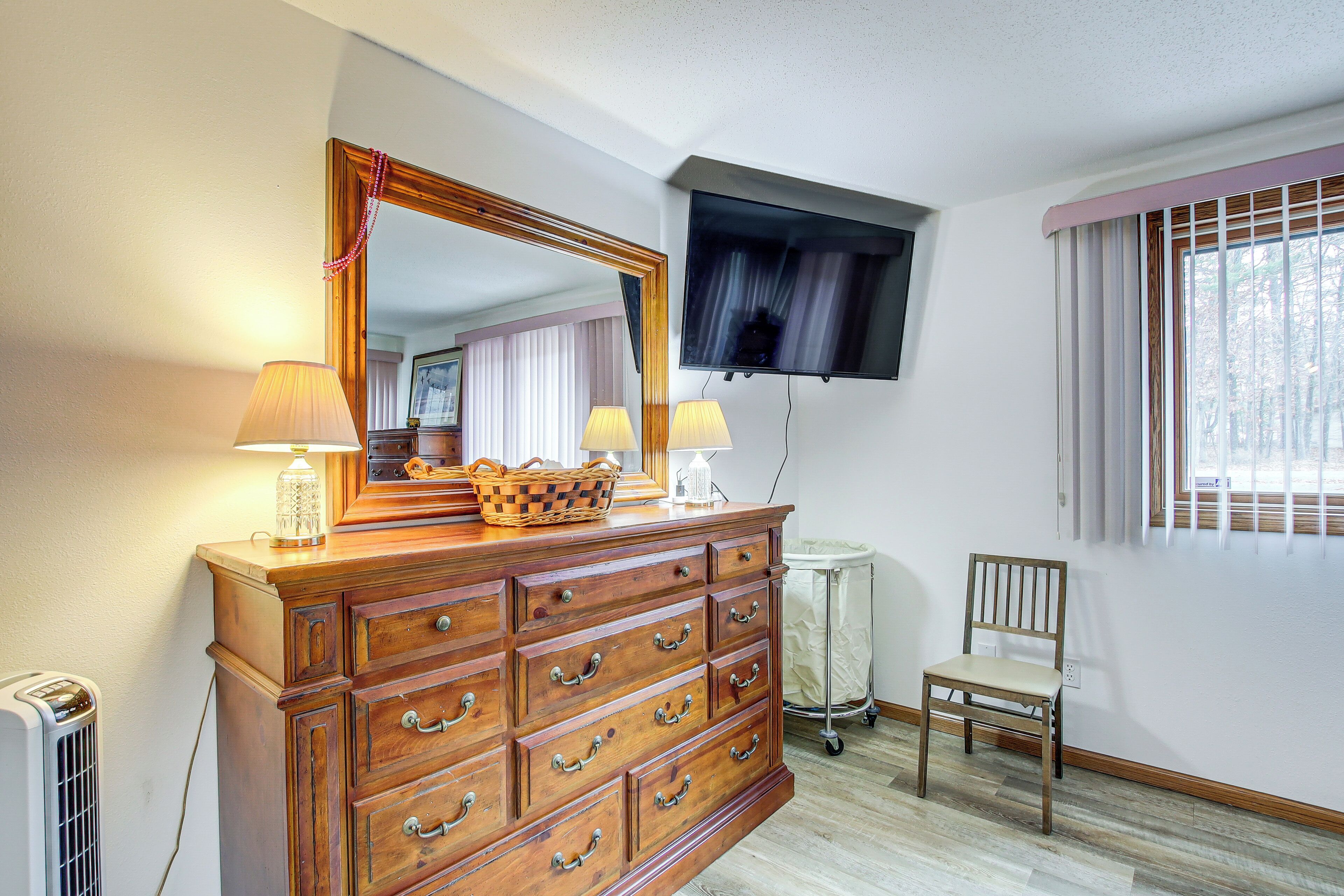Een kamer met een houten dressoir, een spiegel, twee lampen, een televisie, een stoel en een raam met gordijnen.