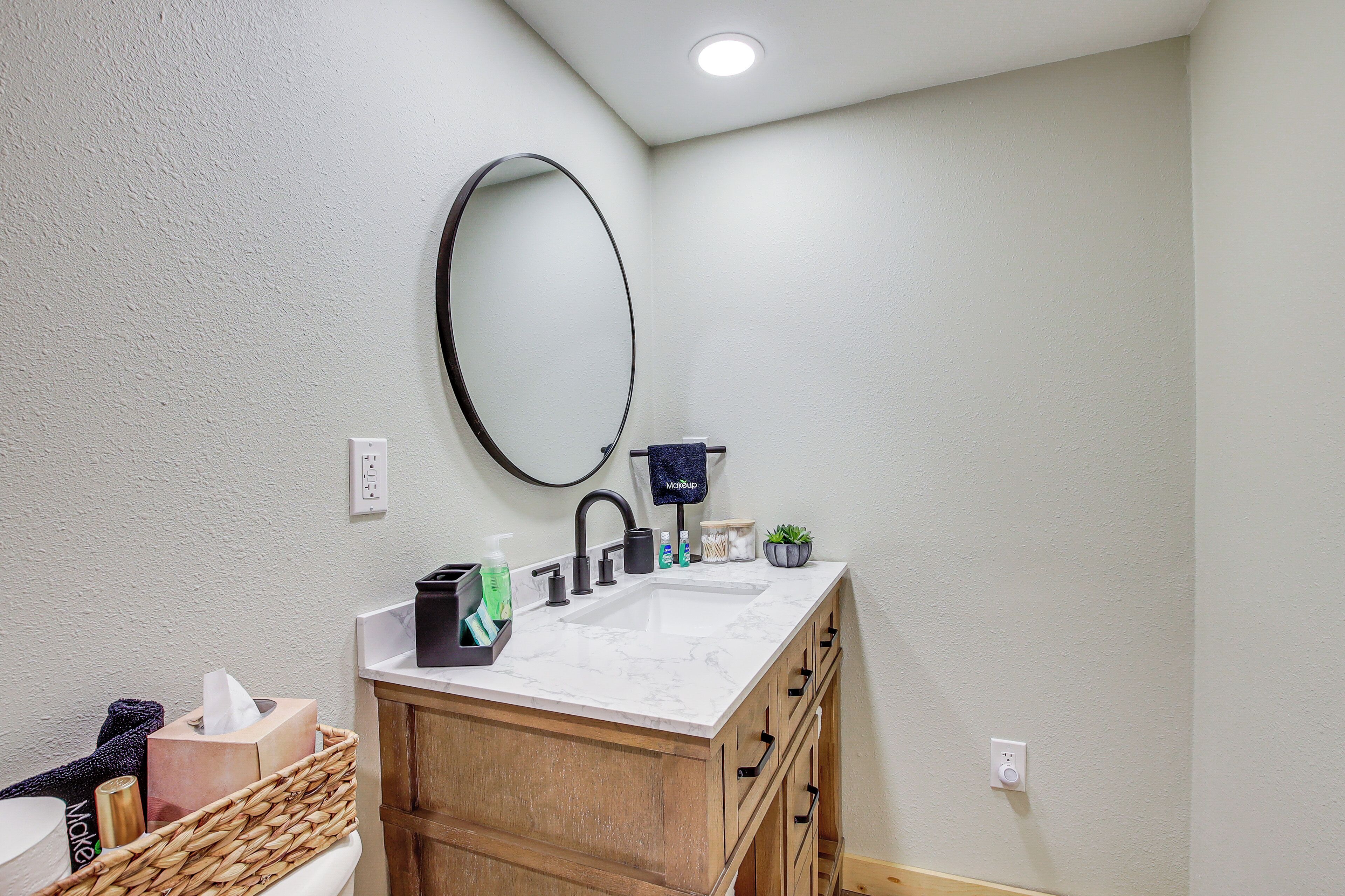 A bathroom with a white marble sink, wooden cabinets, a black faucet, a round mirror, and a small potted plant.