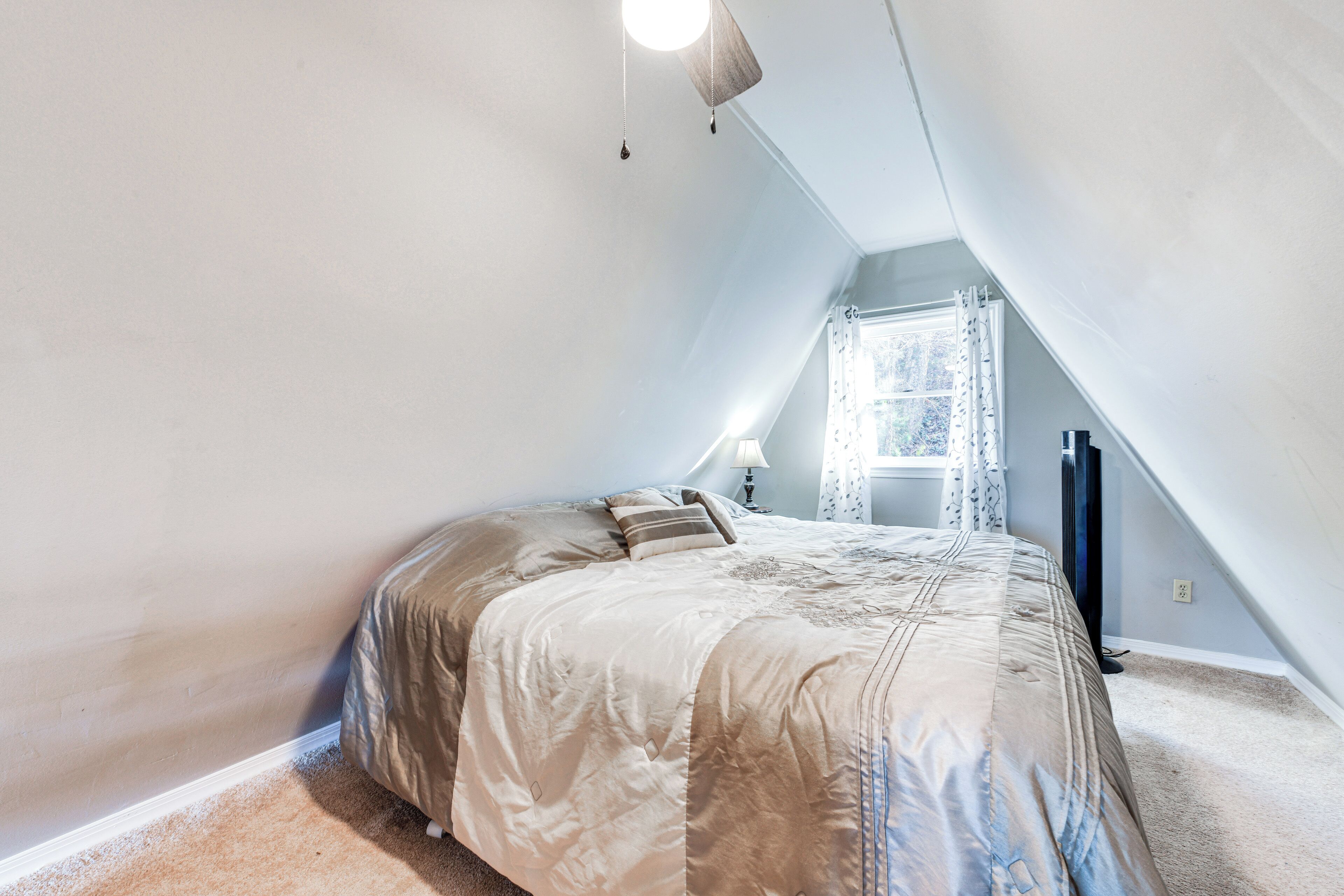 Un dormitorio con techo inclinado, una cama con edredón estampado en tonos beige y azul, una mesita de noche con lámpara y una ventana con cortinas.