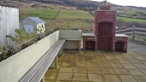 An outdoor patio with a brick oven, a wooden bench, and a white building in the background. - Harefield Holiday Home (Carrick)