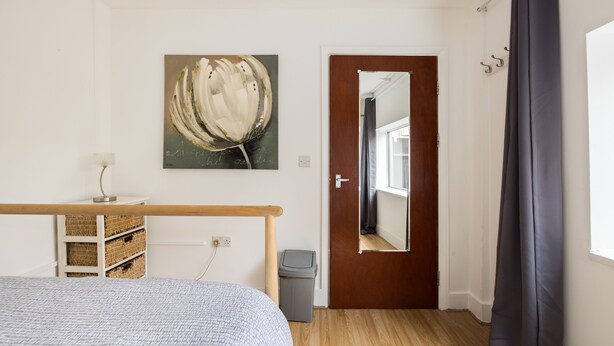 Ein Schlafzimmer mit Bett, Holz-Kopfteil, Wandbild, Tür, Fenster mit Vorhang und einer kleinen Mülltonne.