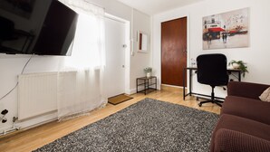 Ein Wohnzimmer mit Flachbildfernseher, braunem Sofa, schwarzem Bürostuhl, einem kleinen Tisch mit Pflanze und einem Bild an der Wand.
