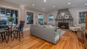 A spacious living room with a stone fireplace, a gray sofa, and a dining area with wooden flooring and chairs. - Lake Winnipesauke compound sleeps 22 in 13 bedrooms stunning sunset views (Alton Bay)