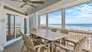 A covered outdoor deck with a table and chairs, large windows offering a view of the beach and ocean.