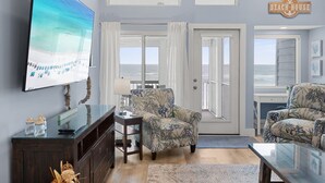 A living room with a large TV, two patterned armchairs, a coffee table, and a view of the ocean through a door.