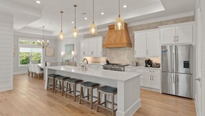 Une cuisine moderne dotée d’un grand îlot central, de tabourets de bar, d’appareils électroménagers en acier inoxydable et de luminaires suspendus.