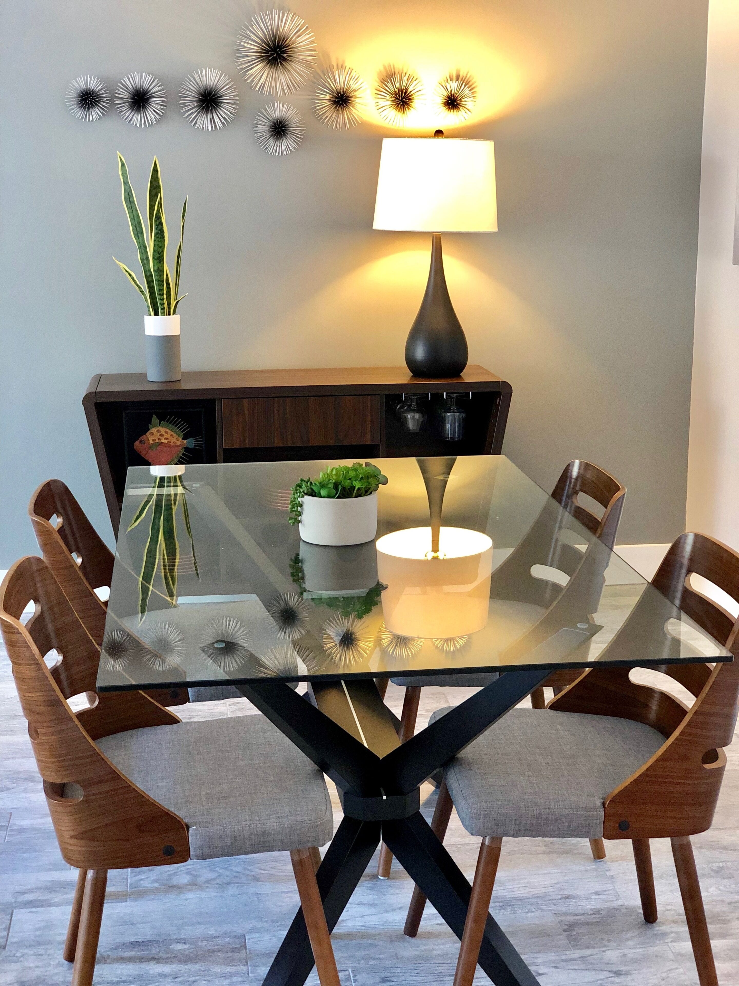Une table à manger avec un plateau en verre, entourée de chaises en bois dotées de coussins en tissu. Une lampe se trouve sur la table, et des éléments décoratifs ornent le mur au-dessus.