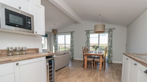 A modern kitchen with white cabinets, a built-in microwave, and a dining area with a wooden table and chairs. - Primrose Lodge (Dartmouth)