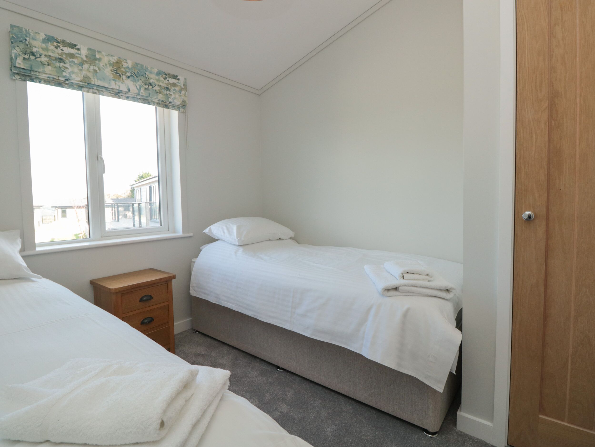 A bedroom with two beds, a window with a view, a wooden nightstand, and a door.