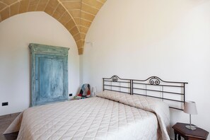 A bedroom with a bed, a nightstand, a lamp, a chair, and a large arched doorway.