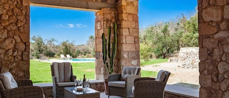 A covered patio with wicker furniture, a stone wall, and a view of a pool and landscape.
