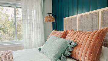 Quarto com cama, roupa de cama branca, almofadas verde-água e laranja, uma luminária trançada e vista para árvores através de uma janela.