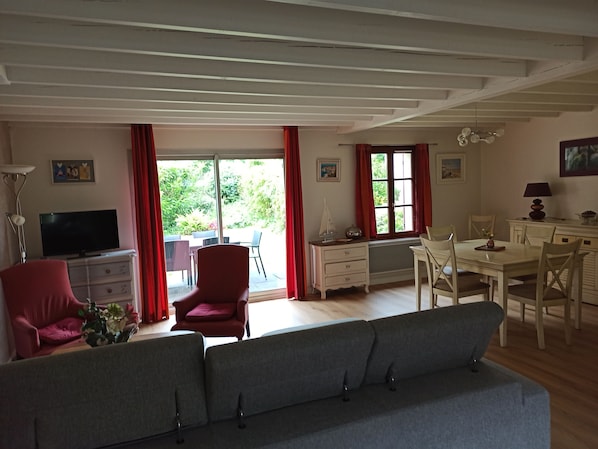 Una sala de estar con sofá, televisión, mesa de comedor y sillas. Hay ventanas con cortinas, un ventilador de techo y una lámpara.
