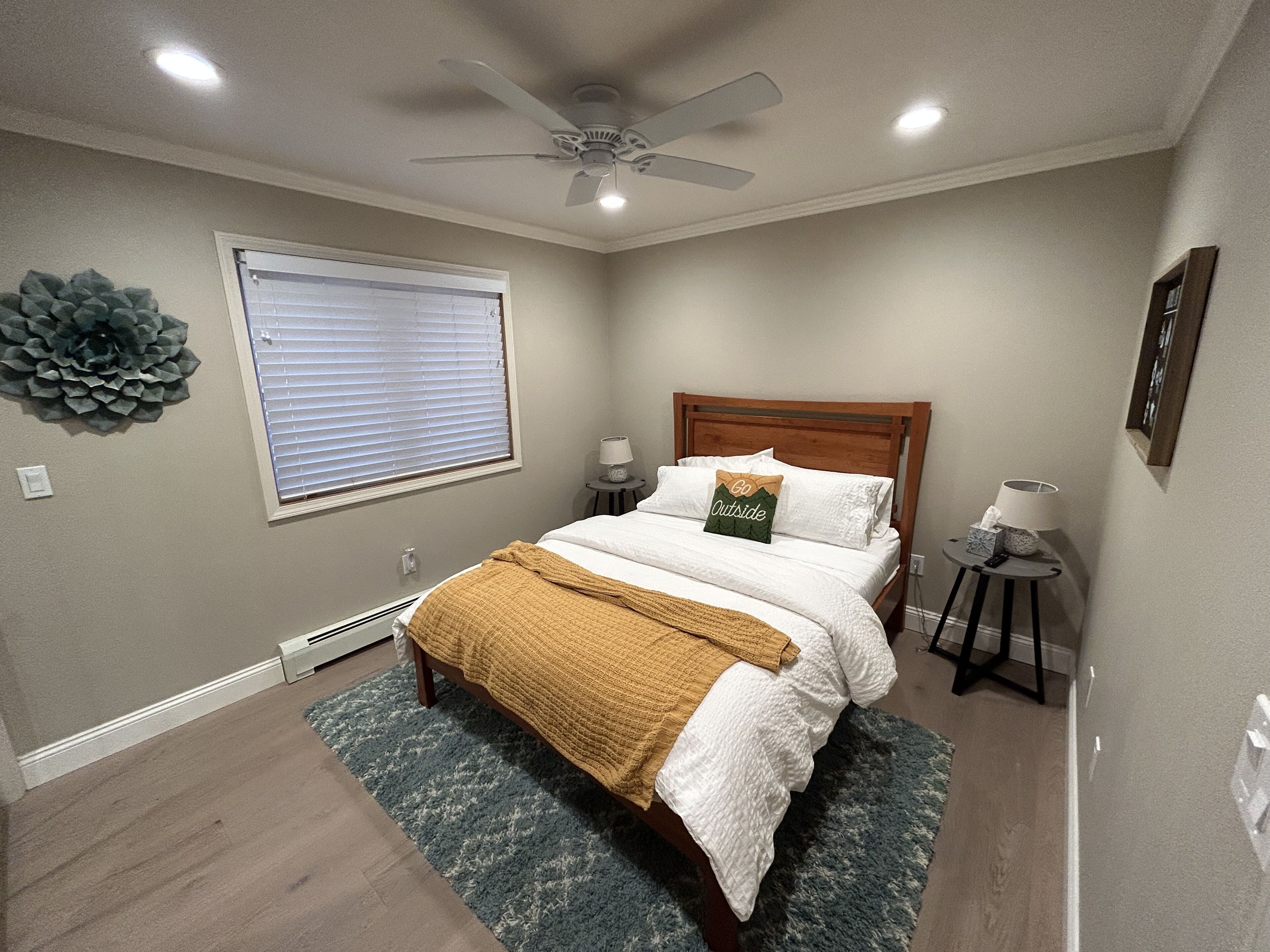 Ein Schlafzimmer mit einem Bett, einem Deckenventilator, einem Fenster mit Jalousien, einem Nachttisch mit Lampe sowie einem dekorativen Wandbild.