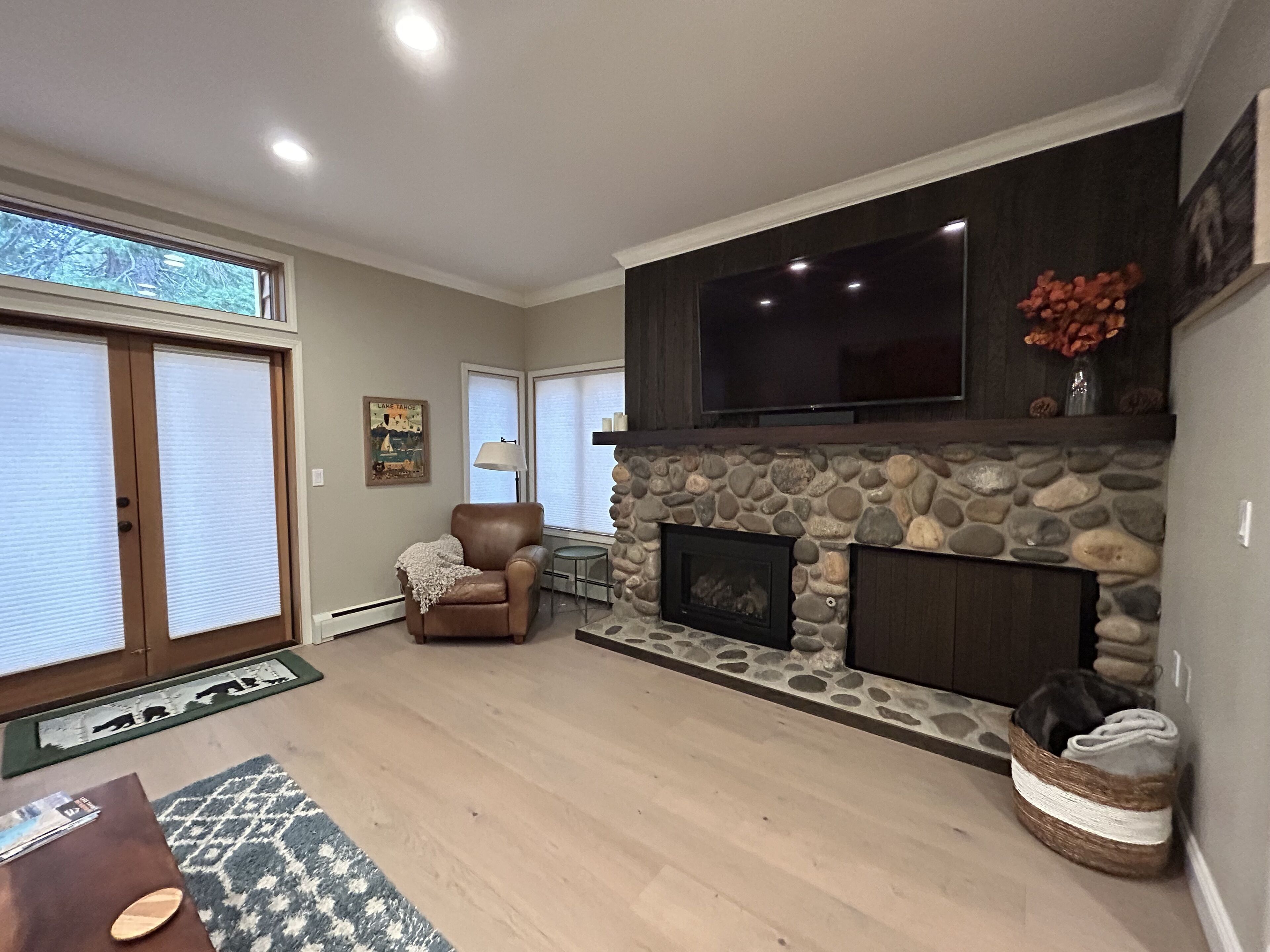 A living room with a stone fireplace, a leather armchair, a television mounted above the fireplace, and a large window with blinds.