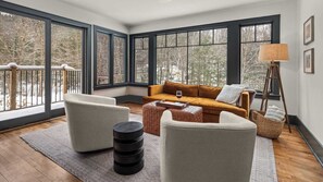 A modern living room with a sofa, two armchairs, a coffee table, and a lamp. Large windows offer a view of trees and a snowy landscape.