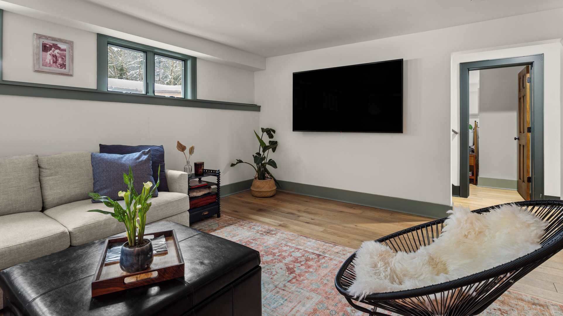 Una sala de estar con un sofá, una mesa de centro con una planta, una silla negra, un televisor en la pared y una puerta que da a otra habitación.