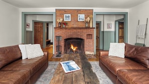 A living room with a brick fireplace, two leather sofas, a coffee table with a book, and framed pictures on the wall. - Spacious 5BR Stowe Resort | Forest & River Views (Stowe)